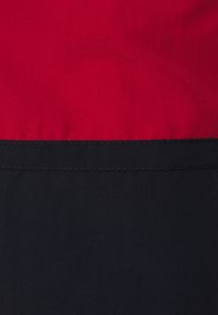 Vörös és fekete textil vízszintesen osztva, sima felülettel; a vörös rész a fekete felett helyezkedik el, egy tiszta varrássor választja el őket.