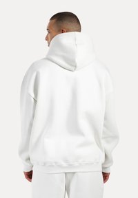Sudadera blanca de gran tamaño con una capucha grande, hombros caídos, puños acanalados y una textura suave y lisa. Sin logotipos ni patrones visibles.