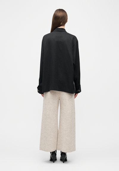 Γυναίκα που στέκεται με την πλάτη στην κάμερα, φορώντας ένα φαρδύ μαύρο πουκάμισο, πλατιά μπεζ πλεκτά παντελόνια και μαύρα γυναικεία μποτίνια με τακούνι, σε λευκό φόντο.