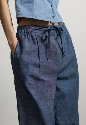 Mujer con pantalones holgados de lino azul oscuro con cordón y una blusa corta azul claro con botones, manos en los bolsillos, sobre fondo liso.