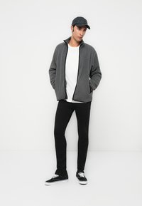 Joven vestido con una chaqueta de forro polar gris con cremallera, una camiseta blanca, pantalones negros ajustados, una gorra negra y zapatillas negras con cordones blancos, de pie con las manos en los bolsillos.