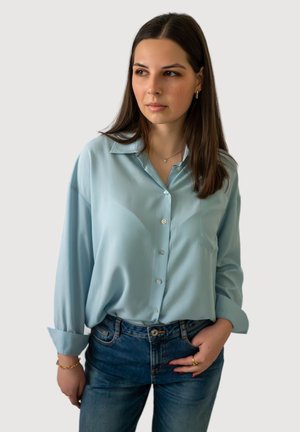 Femme aux longs cheveux bruns portant une chemise à boutons bleu clair et un jean bleu, debout avec une main dans la poche, regardant sur le côté.