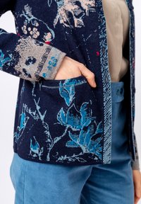 Tumši zila džemperis ar ziedu rakstu zilās un krēmkrāsas nokrāsās, ar kabatu, struktūru audumu un kontrastējošiem adījuma piedurknēm.