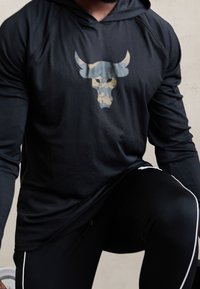 Schwarzer Langarm-Hoodie mit einem Camouflage-Bullenkopf-Graphic auf der Brust. Hat eine entspannte Passform und eine weiche Textur, getragen mit schwarzen Sporthosen.