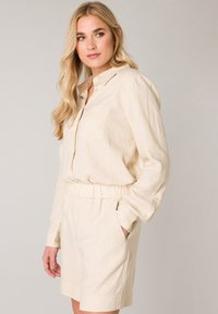Beige linnenshirt jurk met lange mouwen, knoopsluiting aan de voorkant, een enkele borstzak en een elastische taille. Heeft zijzakken en een relaxte pasvorm.