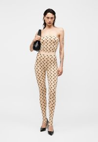 Kobieta ubrana w beżowy kombinezon bez ramiączek z czarnym wzorem półksiężyca, czarne szpilki, niesie czarną torebkę, stoi na białym tle.