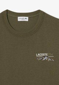 Camiseta de algodón verde oliva con cuello redondo, que presenta el logo "LACOSTE" y un diseño de montaña bordado en blanco y azul. Corte clásico.