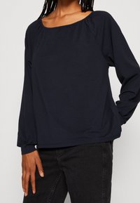 Navy blue long-sleeve top with a wide neckline and gathered accents at the shoulders. Made of soft fabric, featuring a relaxed fit and elastic cuffs.