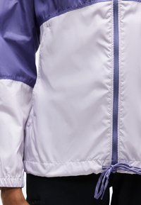 Ανοιχτό μωβ και λεβαντί αδιάβροχο μπουφάν με φερμουάρ μπροστά και ρυθμιζόμενο κορδόνι στο τελείωμα, φορεμένο από άτομο με σκούρο παντελόνι.