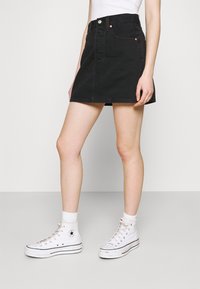 Mujer con minifalda de mezclilla negra, camiseta sin mangas blanca, calcetas blancas y zapatillas altas Converse blancas, de pie frente a un fondo liso.