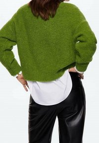 Pull vert court à surface texturée, associé à une chemise blanche en dessous et un pantalon en similicuir noir.