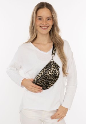 Junge Frau mit langen blonden Haaren, die weiße Kleidung trägt, lächelnd und eine Umhängetasche mit Leopardenmuster quer über der Brust hält.