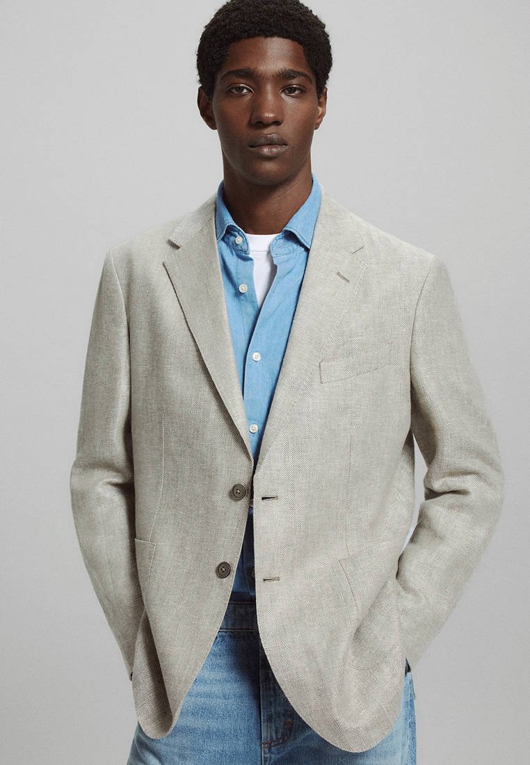 Jeune homme portant un blazer beige clair sur une chemise bleue à boutons et un t-shirt blanc, debout les mains dans les poches devant un fond uni.