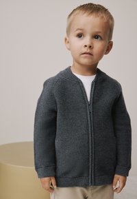Cardigan gris en maille zippé avec texture côtelée, doté d'un col montant et de manches longues. Porté sur une chemise blanche, il complète un pantalon clair.