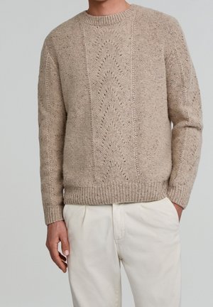 Mand iført beige strikket sweater med chevronmønster og cremefarvede bukser, stående med den ene hånd i lommen mod en ensfarvet baggrund.