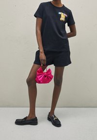 T-shirt nera in cotone con una grafica colorata di un orso, abbinata a pantaloni corti neri e una borsa a forma di fiocco rosa. Le scarpe sono mocassini neri.