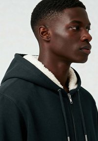 Must mustaga, millel on valge fliisvooder. Omab lukku, tõmblukku ja tekstuurset, pehmet kangast. Suurendatud vaade kaelusele.