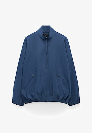 Veste légère bleu marine avec col montant, fermeture éclair frontale, deux poches latérales zippées, poignets et ourlet élastiqués.