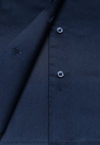 Donkerblauwe, gestructureerde stoffen shirt met knoopsluiting, zichtbare stiksels en twee knoopgaten. Details omvatten een gladde, gelijkmatige afwerking.