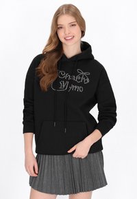 Fekete kapucnis pulóver elülső zsebbel és húzózsinórokkal, strasszköves díszítéssel. Szürke redős szoknyával párosítva.