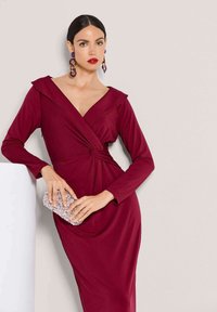 Burdeosfarbenes, langärmliges Kleid mit tiefem V-Ausschnitt und drapiertem Oberteil, mit verdrehtem Taillendetail. In einer Hand wird eine strukturierte silberne Clutch gehalten.