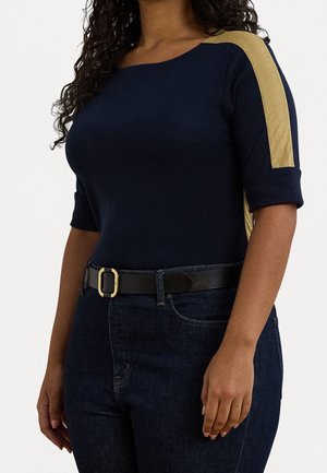 Marineblå top med guldfarvet metallic detalje på skulderen, trekvartlange ærmer, kombineret med mørk denim og et sort bælte med en guldbæltespænde.