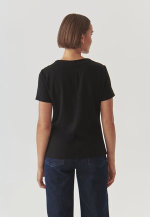 Haut noir à manches courtes avec un col rond et une coupe droite, présentant un détail de couture centrale au dos. Porté avec un pantalon en denim bleu foncé.