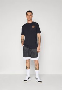 Hombre de pie vistiendo una camiseta gráfica negra, pantalones cortos de baloncesto en blanco y negro, calcetines blancos hasta la mitad de la pantorrilla y zapatillas blancas con detalles en negro.