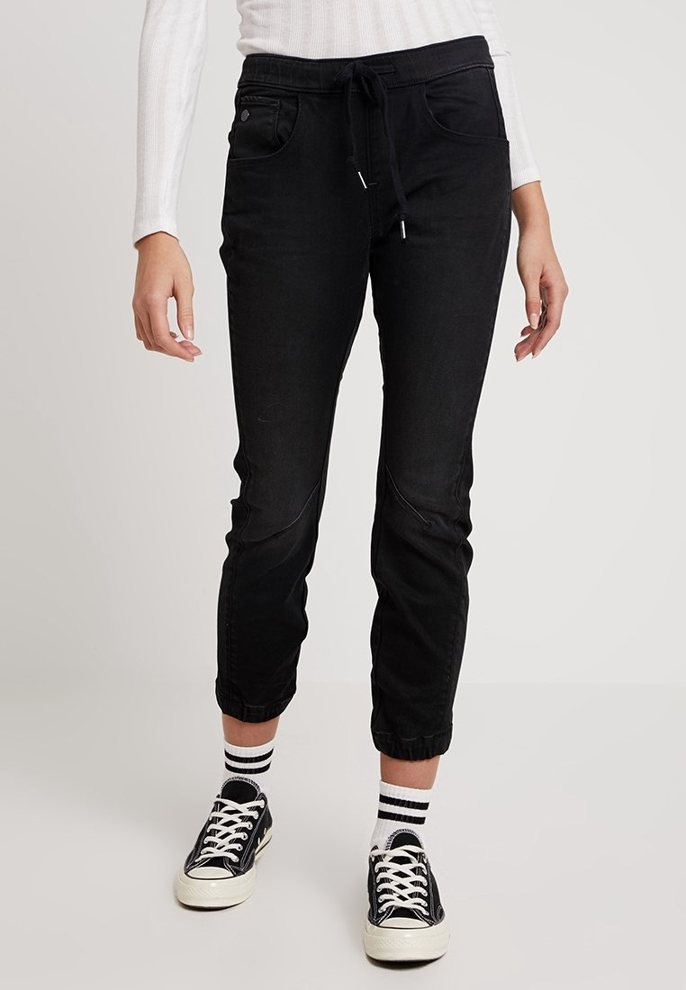 Svarta joggerbyxor med elastiskt midjeband, framfickor, manschetter och en mjuk textur, tillsammans med svart-vita sneakers.