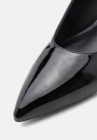 Zapato de tacón alto de charol negro con puntera puntiaguda, textura suave y costuras mínimas. El interior presenta un acabado mate.