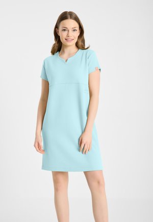 Młoda kobieta uśmiechająca się, ubrana w jasnoniebieską sukienkę o krótkich rękawach i długości do kolan z małym wycięciem przy dekolcie na jednolitym tle.