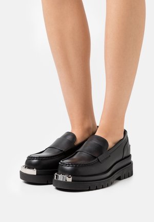 Mocassins en cuir noir à semelles épaisses et plateforme, avec détail en métal argenté, portés pieds nus sur fond blanc.