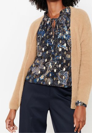 Femme portant un cardigan beige en tissu pelucheux sur un chemisier noir à motifs floraux avec des accents bleus et dorés, associé à un pantalon bleu marine foncé.