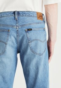Ljust blå denimjeans med rak passform, utrustad med en beige läderbakficka och sydda fickdetaljer.