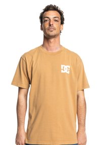 Brown cotton t-shirt with short sleeves, featuring a white DC logo printed on the left chest in a classic design. Smooth texture.