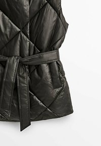 Sort quiltet vest med bælte i taljen, der har en glat, skinnende overflade og diagonale syninger. Inkluderer sidelommer.