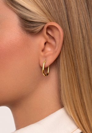 Pendiente de aro dorado con forma suave y redondeada. La superficie tiene una textura pulida que refleja la luz, llevado en la oreja de un modelo.