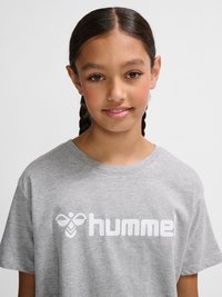 Junge Person mit geflochtenem Haar, die ein graues T-Shirt mit weißem „hummel“-Logo auf einem einfarbigen hellen Hintergrund trägt.