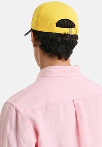 Roze linnen overhemd met een rugplooitje en een korte kraag; een gele pet met een verstelbare band zit op korte, donkere krullend haar.