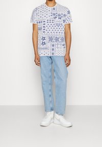 Valkoinen lyhythihainen t-paita, jossa on monimutkaisia sinisiä kuvioita, yhdistettynä vaaleansinisiin farkkuihin ja valkoisiin lenkkareihin. Tyylikäs muotoilu ja rento istuvuus.