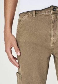 Pantalons cargo marron en tissu texturé, avec des poches latérales et des coutures visibles. Le tissu a un léger brillant, ce qui améliore son apparence.