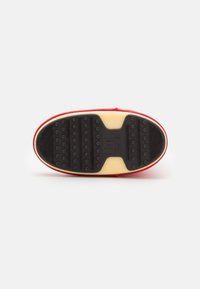 Sole of a red shoe with black rubber treads featuring round grips and ridges, on a beige background, viewed from bottom-up.
