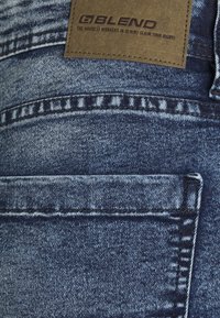 Džínové kalhoty v tmavě modré barvě se slabě opotřebovanou texturou. Mají koženou nášivku s logem a přední kapsu s viditelným švem a nitěmi.