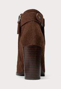 Stivale alla caviglia in suede marrone con punta arrotondata, zip laterale e tacco in legno curvo. Presenta una fascia con dettaglio a bottone alla caviglia.