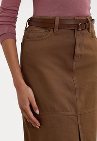 Kvinna iklädd en åtsittande brun kjol med framficka, matchande brun bälte och en mauve långärmad topp, med handen vilande vid sidan.