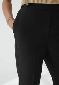 Zwarte op maat gemaakte broeken met een gladde textuur, voorzien van zijzakken en een knoopaccent aan de taille, gezien vanaf de zijkant met een gebreide top.