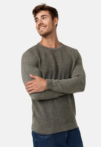 Grå strikket sweater med et struktureret mønster, rund halsudskæring og kontrasterende kanter, parret med blå jeans. Central figur med korslagte arme.