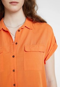 Pomarańczowa bluzka z krótkim rękawem wykonana z lekkiego materiału, z zapięciem na guziki z przodu, dwoma kieszeniami na piersi i luźnym krojem.