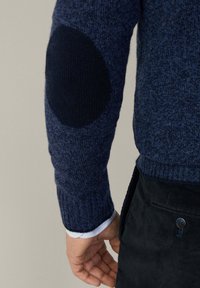 Maglione blu navy lavorato a maglia con polsini a coste e finitura testurizzata. Presenta un toppe circolare scura sul gomito. Una mano si avvicina a un pantalone in velluto a coste scuro.