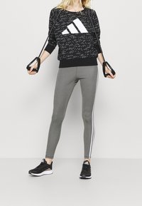 Sudadera negra con manchas blancas y gran logo blanco, combinada con mallas gris ajustadas que tienen una franja lateral blanca. Zapatillas deportivas negras.